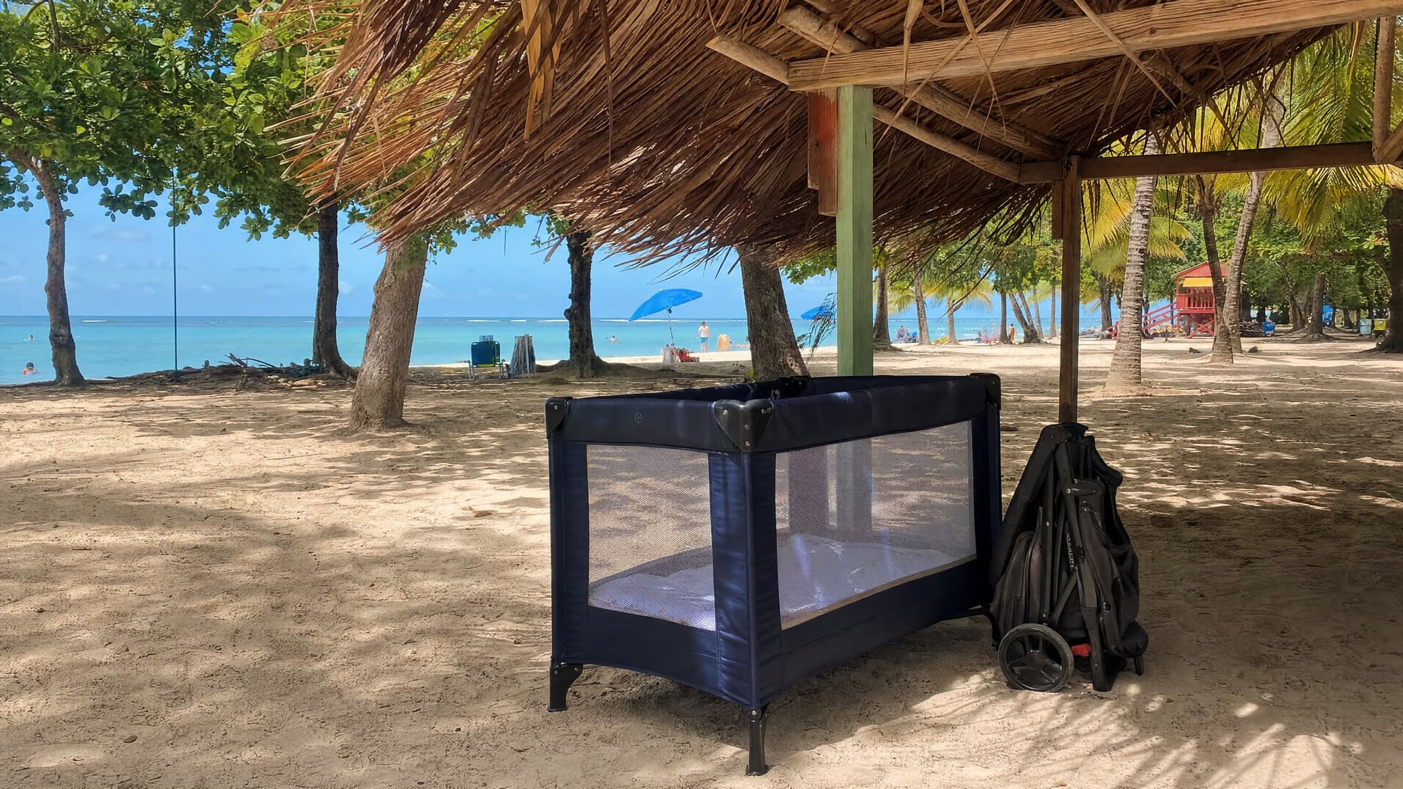 A travel crib and folded stroller set up in the shade of a beach palapa, ready for a family beach day